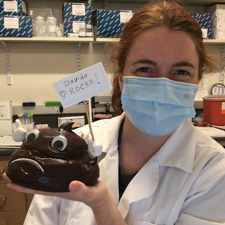 Davida Smyth wearing a mask in a lab holding a sign that says "Davida Rocks" along with a anthropomorphic poop figurine. 